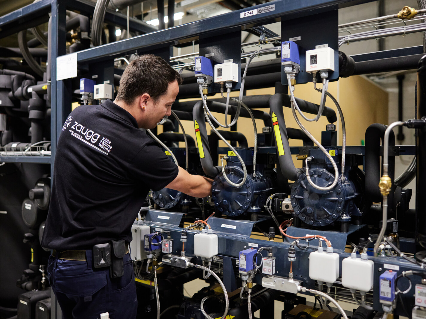 Ein Mitarbeiter der f. zaugg ag arbeitet an einer komplexen CO2 Kälteanlage. Er trägt ein schwarzes Polohemd mit dem Firmenlogo und führt technische Einstellungen an den Kompressoren und Leitungen der Anlage durch. Die Umgebung zeigt ein industrielles Umfeld mit zahlreichen Kabeln, Rohren und Komponenten.