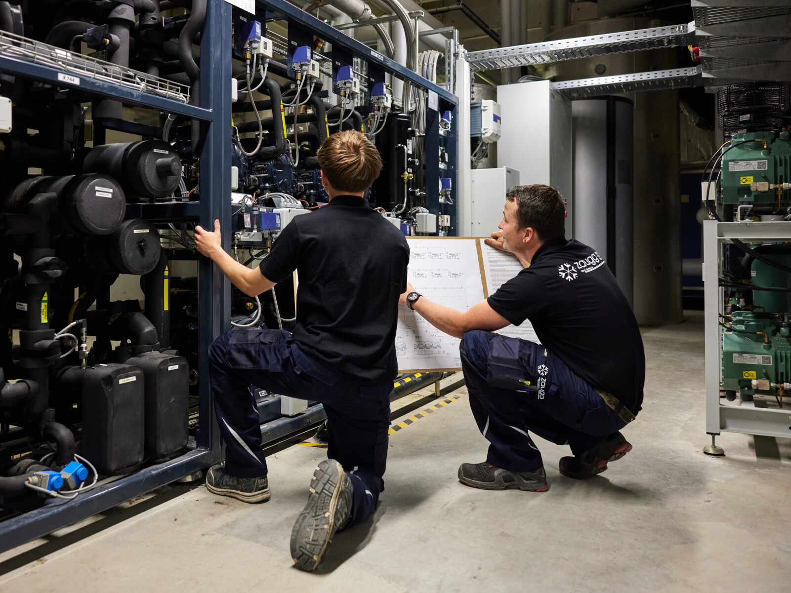 Ein Kältesystemmonteur schaut sich mit einem Lernenden eine Kälteanlage an und erklärt die Kühlanlage. Professionelle Kältetechnik Schweiz.